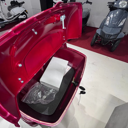 Izdelek je električni skuter s tremi kolesi, model Vigorous Vista 1. Na sliki je prikazan notranji prostor prtljažnika, ki je rdeče barve. V prtljažniku se nahajajo škatla in plastična vrečka, kar nakazuje na možnost shranjevanja osebnih predmetov. V ozadju so vidni tudi drugi električni skuterji.