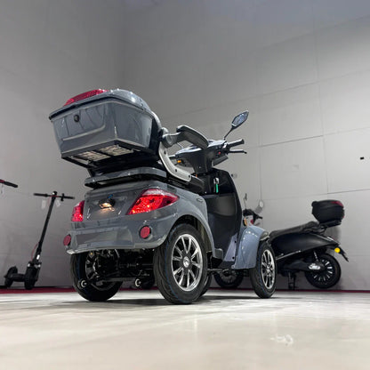 Izdelek na sliki je električni mobilni skuter, model Vigorous Vista Q1. Na sliki je vidna zadnja stran skuterja, ki ima štiri kolesa, prostoren prtljažnik na zadnjem delu ter udobno sedežno površino. Skuter je zasnovan za enostavno manevriranje in udobno vožnjo, kar ga naredi primernega za različne uporabnike. V ozadju so vidni tudi drugi električni skuterji.