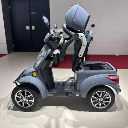 Izdelek na sliki je električni mobilni skuter, model Vigorous Vista Q1. Na sliki je prikazan skuter s štirimi kolesi, ki ima prostorno sedežno mesto in prtljažnik. Barva skuterja je siva, opremljen je z ogledali in ima tudi zaščitno čelado na zadnjem delu. V ozadju so vidni drugi skuterji.