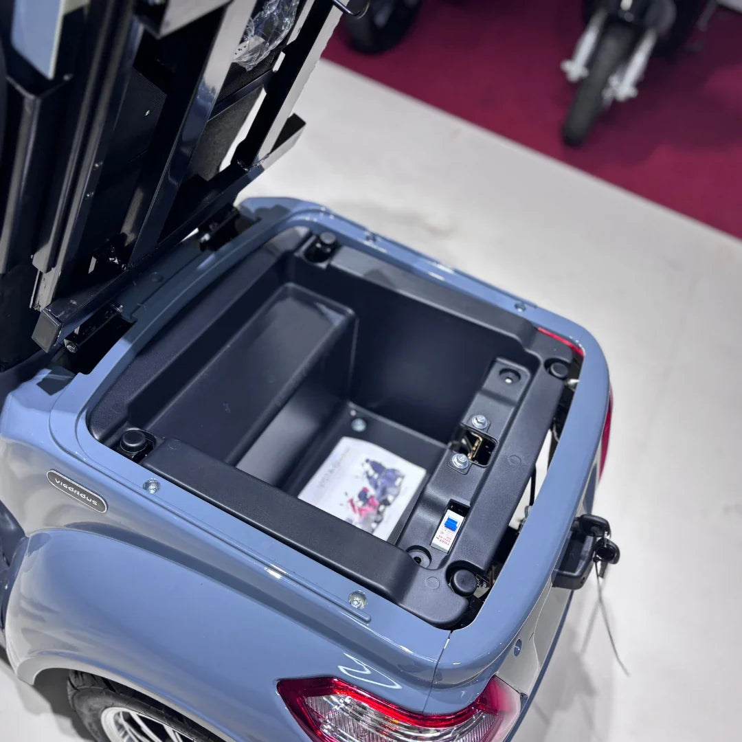 Izdelek na sliki je električni mobilni skuter, model Vigorous Vista Q1. Na sliki je vidna odprta predalna enota, ki razkriva prostor za shranjevanje, verjetno namenjen bateriji in dodatnim predmetom. Barva skuterja je svetlo modra, kar mu daje sodoben videz. V ozadju so vidni tudi drugi skuterji, kar nakazuje, da je slika posneta v prodajnem prostoru.