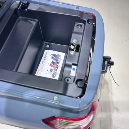 Izdelek je električni mobilni skuter, natančneje štirikolesni električni skuter model Vigorous Vista Q1. Na sliki je prikazan prostor za shranjevanje, ki se nahaja na zadnjem delu skuterja. V notranjosti je vidna oznaka z informacijami o modelu, ki vključuje slike različnih barv skuterja. Skuter ima moder plastični pokrov in je zasnovan za udobno vožnjo.