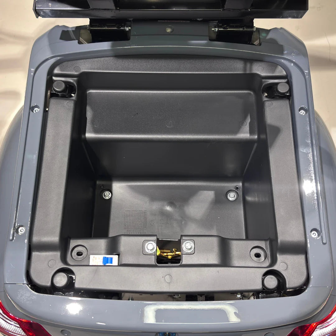 Izdelek je električni mobilni skuter, model Vigorous Vista Q1. Na sliki je prikazan odprt prostor za shranjevanje, ki se nahaja na zadnjem delu skuterja. V notranjosti je vidna plastična struktura brez dodatnih predmetov, kar nakazuje, da je prostor pripravljen za shranjevanje.