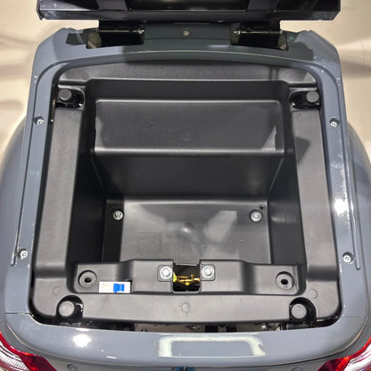 Izdelek je električni mobilni skuter, model Vigorous Vista Q1. Na sliki je prikazan odprt prostor za shranjevanje, ki se nahaja na zadnjem delu skuterja. V notranjosti je vidna plastična struktura brez dodatnih predmetov, kar nakazuje, da je prostor pripravljen za shranjevanje.