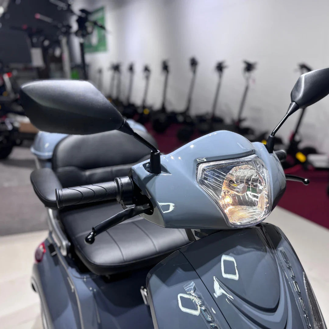 Izdelek na sliki je električni skuter, natančneje model Vigorous Vista Q1. Na sliki je prikazan sprednji del skuterja, kjer so vidni krmilo, smerniki in luč. V ozadju so vidni tudi drugi električni skuterji, kar nakazuje, da gre za razstavo ali trgovino z električnimi vozili. Skuter ima udobno sedežno površino in ergonomsko zasnovano krmilo.