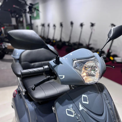 Izdelek na sliki je električni skuter, natančneje model Vigorous Vista Q1. Na sliki je prikazan sprednji del skuterja, kjer so vidni krmilo, smerniki in luč. V ozadju so vidni tudi drugi električni skuterji, kar nakazuje, da gre za razstavo ali trgovino z električnimi vozili. Skuter ima udobno sedežno površino in ergonomsko zasnovano krmilo.