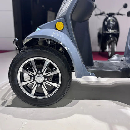 Izdelek je električni mobilni skuter, model Vigorous Vista Q1. Na sliki je prikazan del skuterja, ki vključuje sprednje kolo z aluminijastim platiščem in široko pnevmatiko. Vidna je tudi svetilka na sprednjem delu in del talne površine, kar kaže na zasnovo, ki je prilagojena udobni vožnji. V ozadju je še en skuter, kar nakazuje, da je slika posneta v razstavnem prostoru.