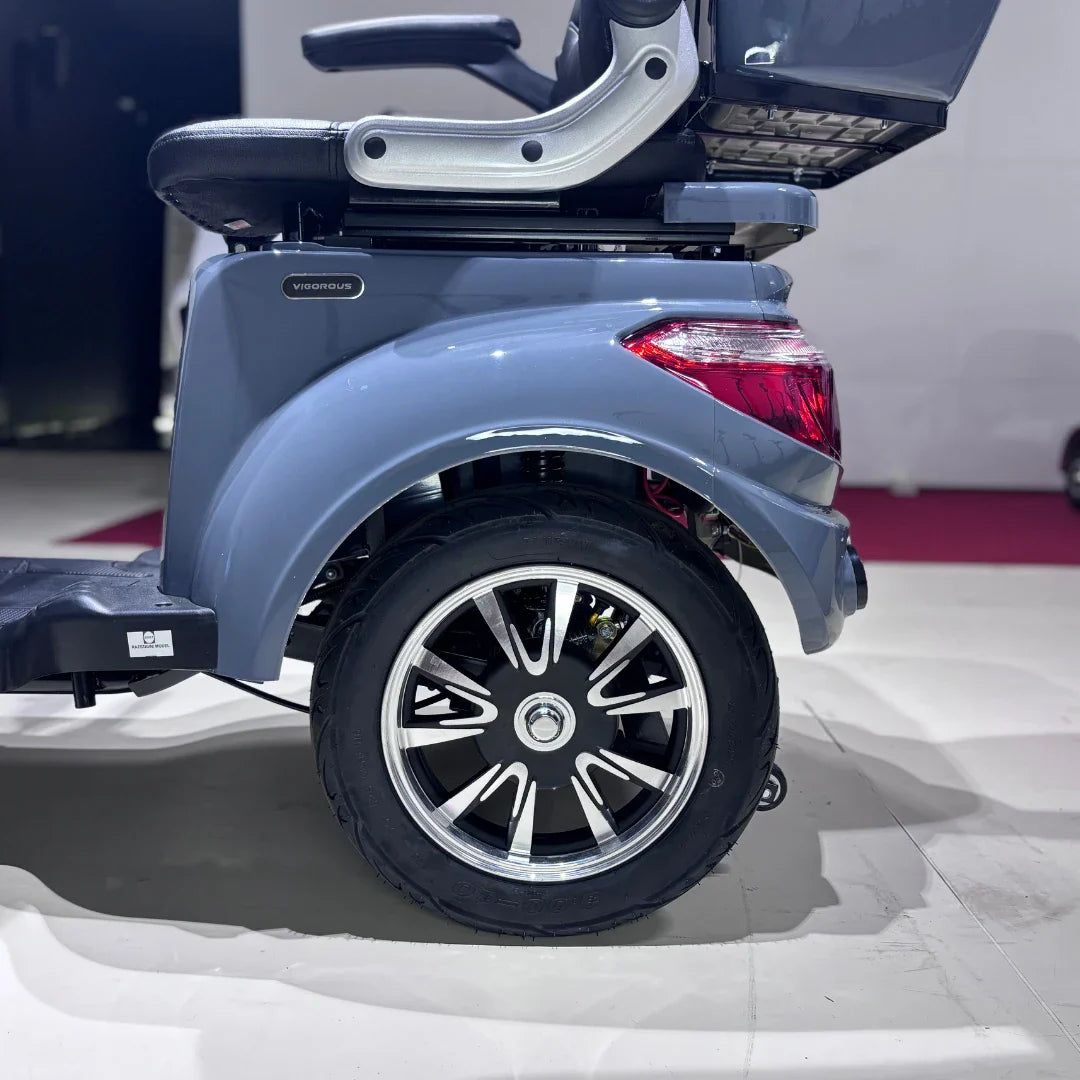 Izdelek je električni mobilni skuter, model Vigorous Vista Q1. Na sliki je prikazan del skuterja, ki vključuje zadnje kolo s športnim dizajnom platišča in del karoserije v svetlo sivi barvi. Vidna je tudi udobna sedežna garnitura ter del podvozja. Skuter je zasnovan za udobno in enostavno premikanje.