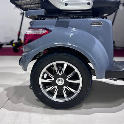 Izdelek je električni mobilni skuter, model Vigorous Vista Q1. Na sliki je prikazan zadnji del skuterja, kjer je vidna kromirana platišča in črn pnevmatik. Skuter ima robustno zasnovo, kar omogoča udobno vožnjo. V ozadju so vidni tudi drugi električni skuterji.