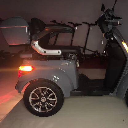 Izdelek je električni skuter, natančneje štirikolni električni mobilni skuter, model Vigorous Vista Q1. Na sliki je prikazana stranska perspektiva skuterja, ki ima udoben sedež in prostoren prtljažnik. Vidni so tudi detajli na kolesu ter zadnja luč, kar kaže na funkcionalnost in obliko skuterja. V ozadju so vidni tudi drugi električni skuterji.