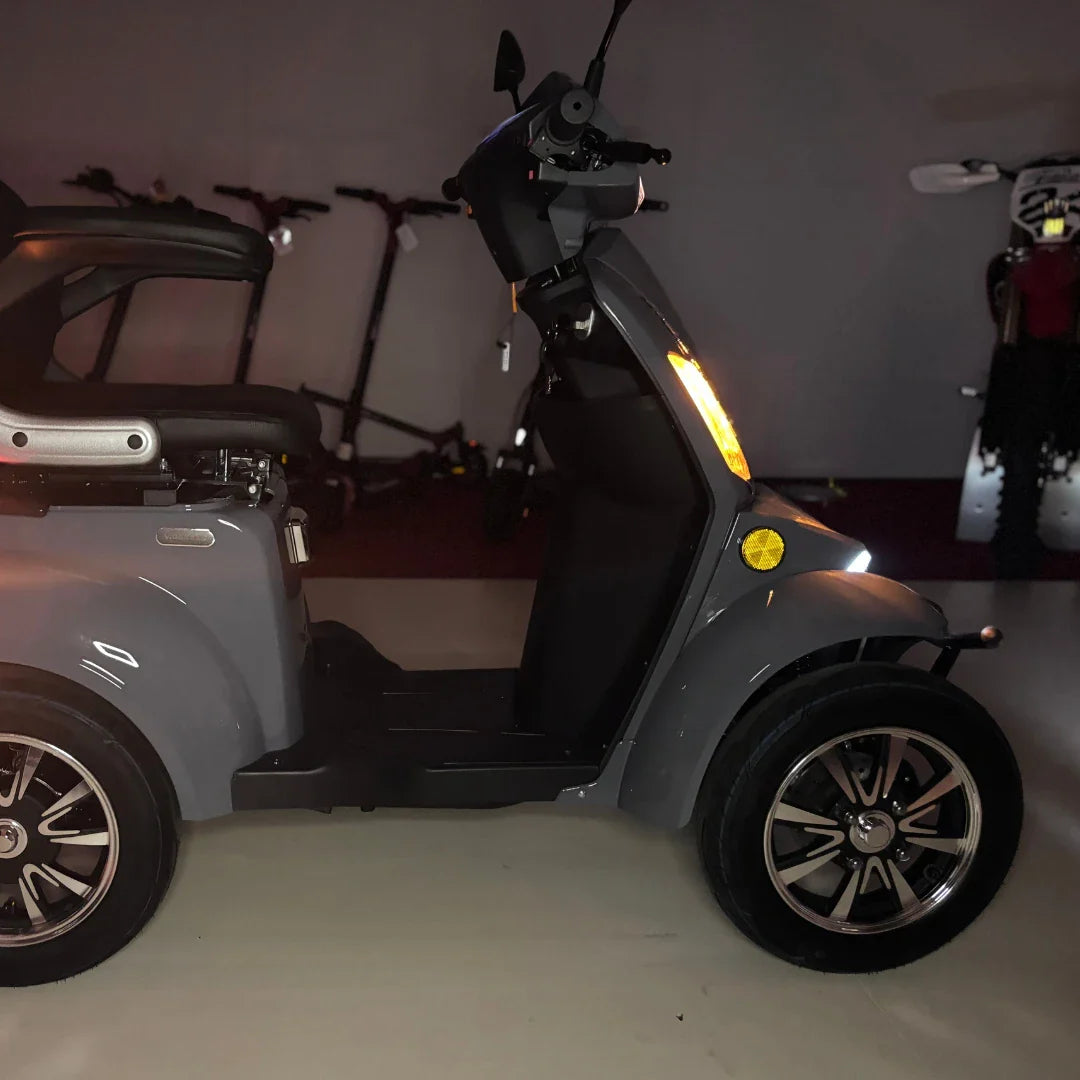 Izdelek na sliki je električni mobilni skuter, model Vigorous Vista Q1. Na sliki je vidna stranska perspektiva skuterja, ki ima tri kolesa in udobno sedišče. Opremljen je z LED lučmi in ima sodoben dizajn. V ozadju so vidni tudi drugi električni skiroji, kar nakazuje, da gre za razstavo ali prodajni prostor.