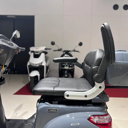 Izdelek je električni mobilni skuter, model Vigorous Vista Q1. Na sliki je vidna udobna sedežna enota s naslonjalom in ročaji, ki zagotavljajo podporo med vožnjo. V ozadju so vidni tudi drugi skuterji, kar nakazuje, da gre za prodajno mesto ali razstavo. Sedež je oblazinjen in zasnovan za udobno vožnjo.