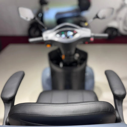 Izdelek je električni mobilni skuter, model Vigorous Vista Q1. Na sliki je prikazan pogled na sedež in krmilo skuterja. Sedež je oblazinjen in ima naslonjala za roke, medtem ko je na krmilu vidna nadzorna plošča z različnimi kontrolami in indikatorji. Skuter je zasnovan za udobno in enostavno vožnjo.