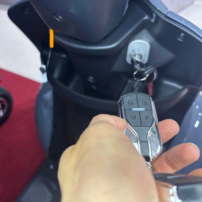 Izdelek je električni mobilni skuter, model Vigorous Vista Q1. Na sliki je vidna roka, ki drži daljinski upravljalnik, ki ima več gumbov, ter ključavnica, vgrajena v skuter. V ozadju je del konstrukcije skuterja, kar nakazuje na njegovo zasnovo in funkcionalnost.