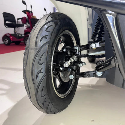 Izdelek je štirikolesni električni mobilni skuter, model Vigorous Vista Q1. Na sliki je prikazan del kolesa, ki vključuje pnevmatiko z izrazitim profilom in del mehanizma vzmetenja. V ozadju so vidni še drugi mobilni skuterji. Pnevmatika je zasnovana za dobro oprijemljivost in stabilnost med vožnjo.