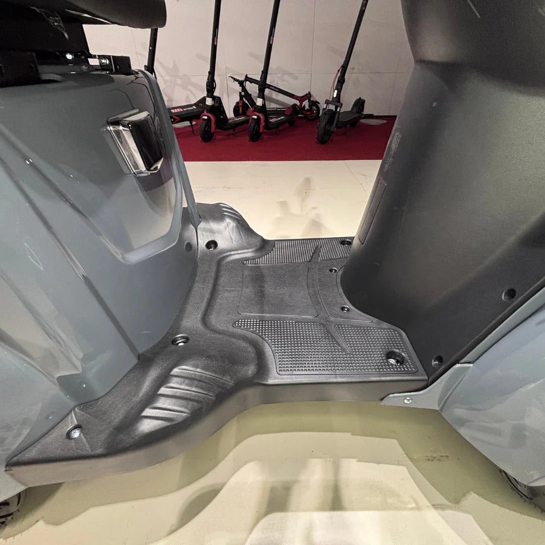 Izdelek je električni mobilni skuter, model Vigorous Vista Q1. Na sliki je prikazan del podnožja skuterja, ki ima teksturirano površino za boljši oprijem. V ozadju so vidni tudi drugi električni skiroji, kar nakazuje, da gre za okolje, kjer se ponujajo različni električni prevozi.