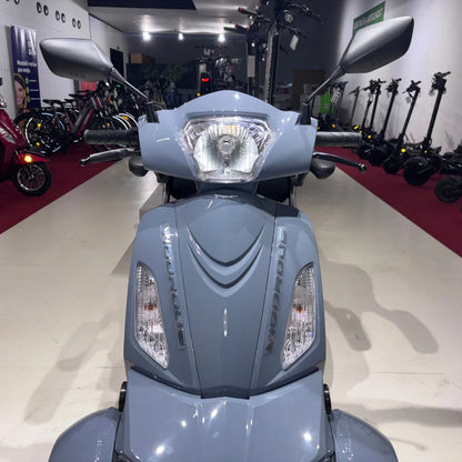 Izdelek na sliki je električni mobilni skuter, model Vigorous Vista Q1. Na sliki je vidna sprednja stran skuterja, ki ima moderno obliko, svetlo središče in dva stranska ogledala. V ozadju so vidni tudi drugi električni skuterji in kolesa, kar nakazuje, da gre za trgovino ali razstavo električnih vozil. Skuter je v svetlo sivi barvi.