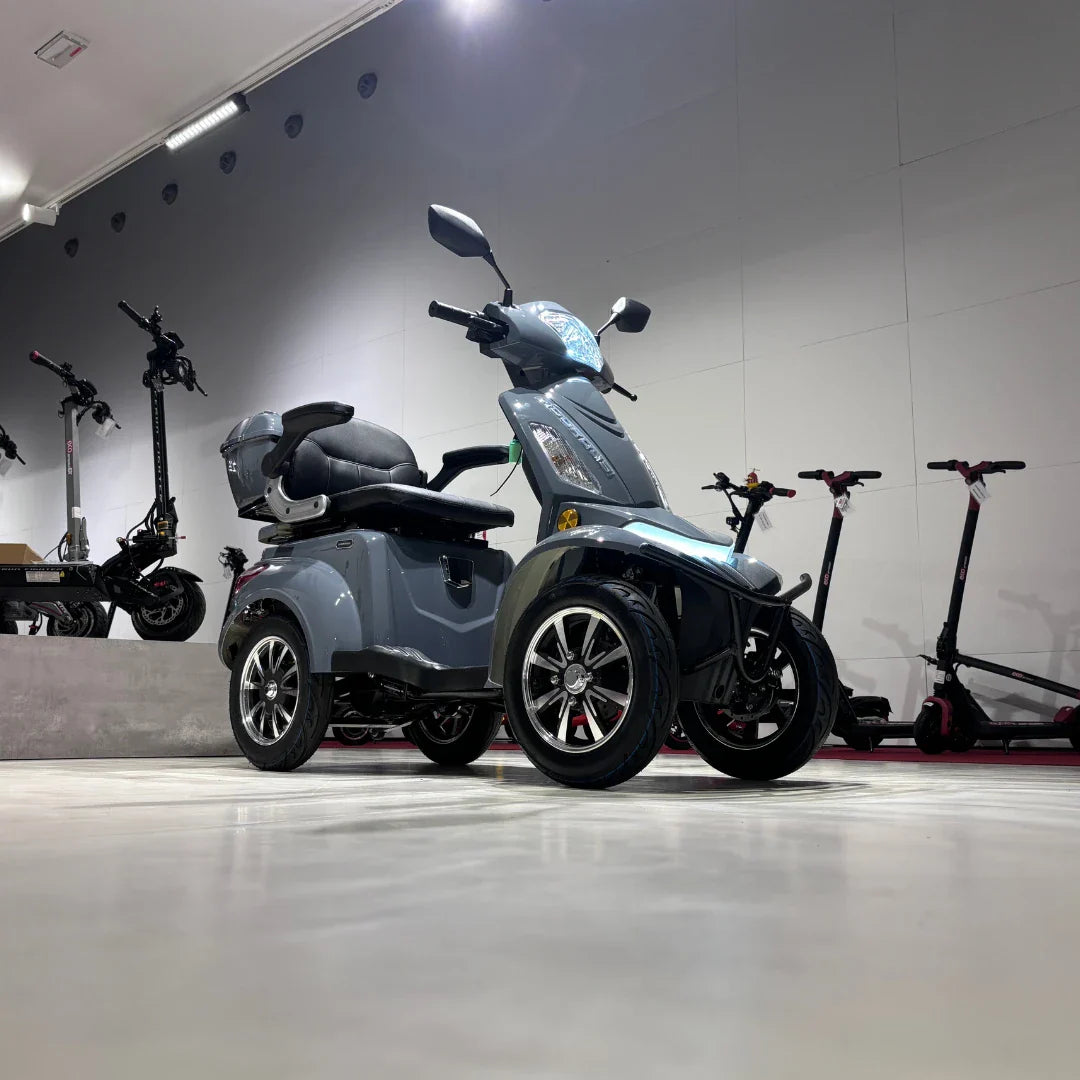 Izdelek na sliki je električni mobilni skuter, model Vigorous Vista Q1. Skuter ima tri kolesa, kar zagotavlja stabilnost in udobno vožnjo. Na sliki je vidna moderna zasnova s širokim sedežem in udobnim naslonjalom. V ozadju so vidni tudi električni skiroji, kar nakazuje, da gre za prodajno ali razstavno okolje.