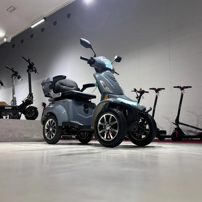 Izdelek na sliki je električni mobilni skuter, model Vigorous Vista Q1. Skuter ima tri kolesa, kar zagotavlja stabilnost in udobno vožnjo. Na sliki je vidna moderna zasnova s širokim sedežem in udobnim naslonjalom. V ozadju so vidni tudi električni skiroji, kar nakazuje, da gre za prodajno ali razstavno okolje.