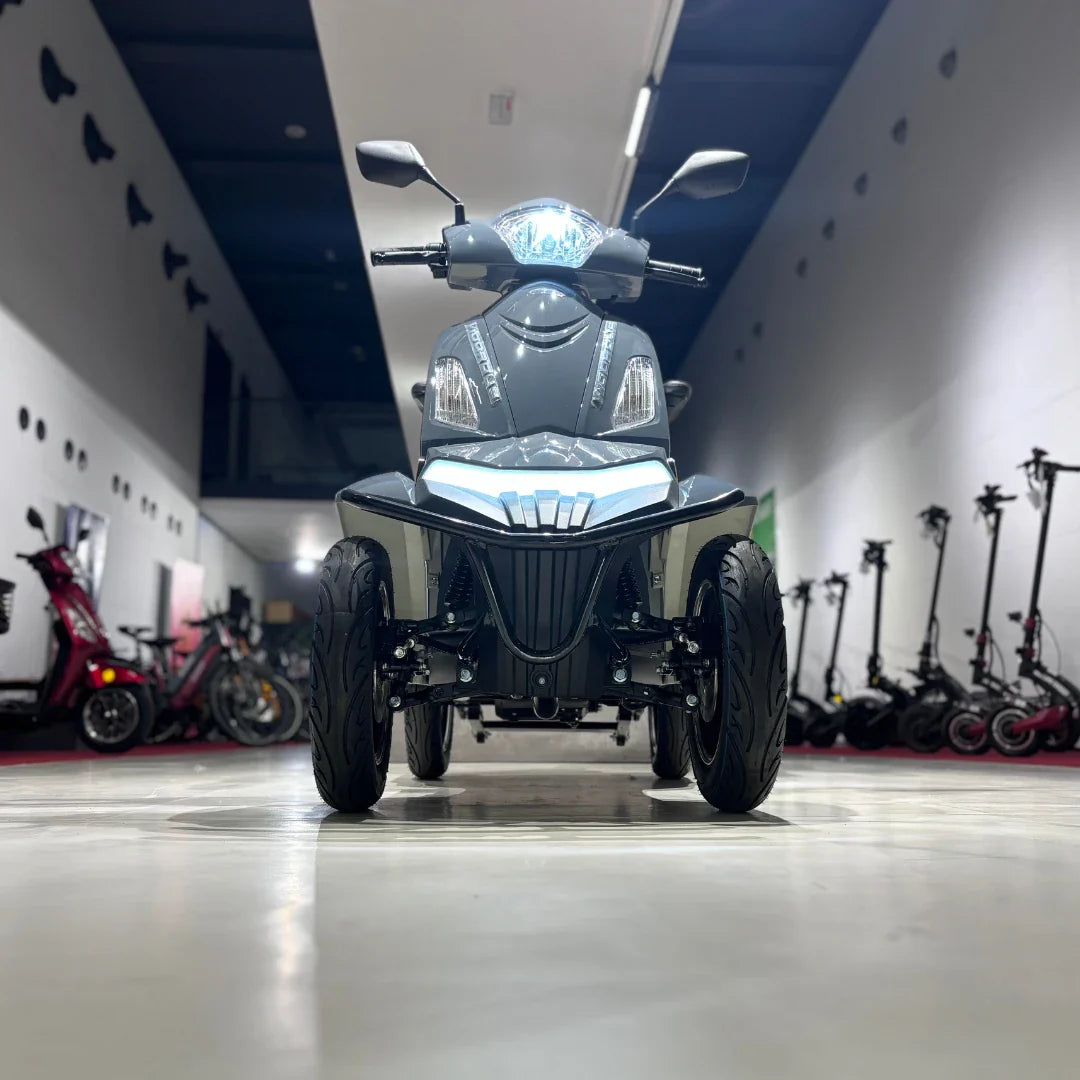 Izdelek je električni mobilni skuter, model Vigorous Vista Q1. Na sliki je prikazan sprednji del skuterja, ki ima moderno obliko, LED luči in široke pnevmatike. V ozadju so vidni tudi drugi električni skuterji, kar nakazuje na okolje, kjer so shranjeni ali razstavljeni. Skuter je zasnovan za udobno in varno vožnjo.