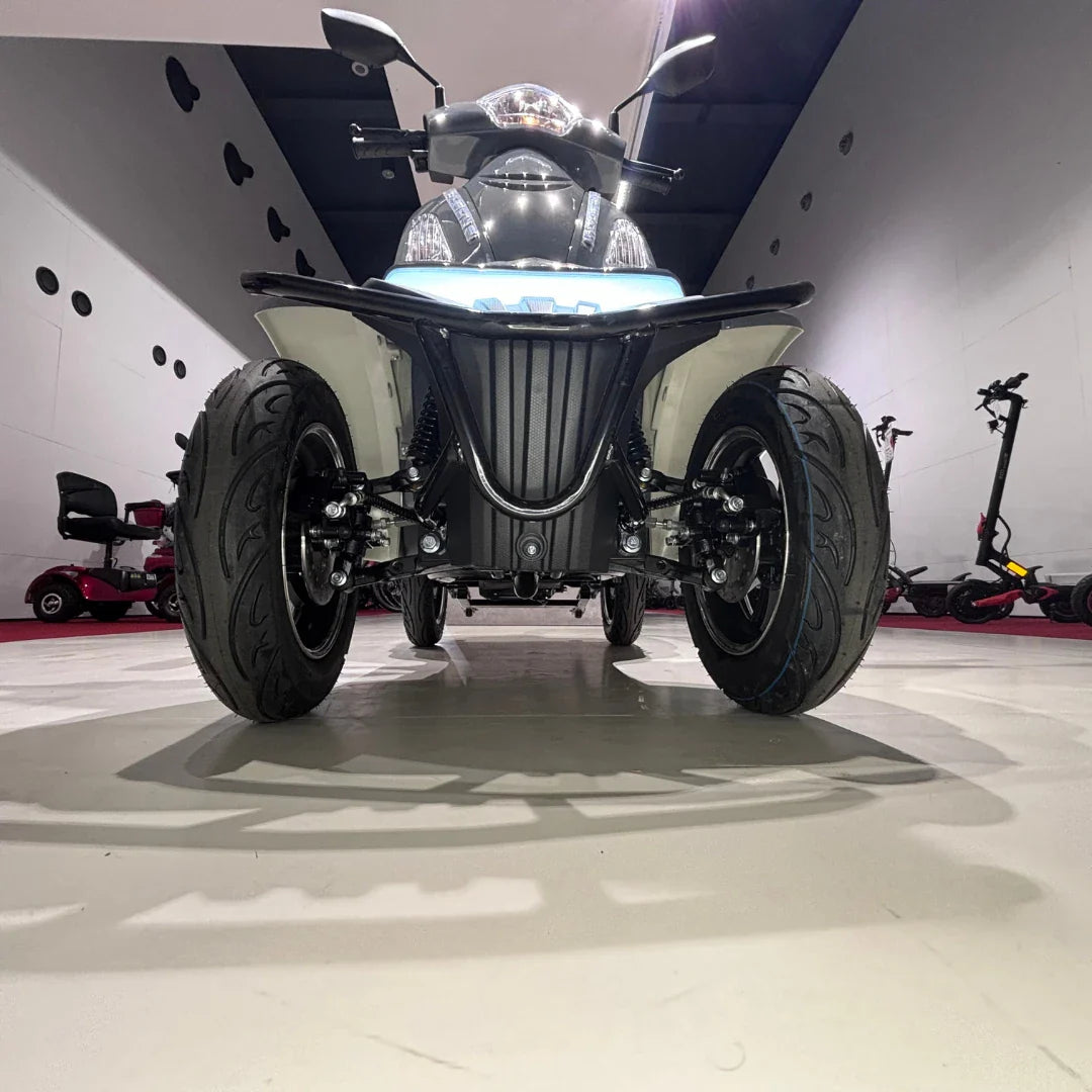 Izdelek na sliki je električni mobilni skuter, specifično model Vigorous Vista Q1. Na sliki je prikazan sprednji del skuterja, ki ima štiri široke pnevmatike, kar omogoča stabilno vožnjo. Vidne so tudi luči in krmilo, kar nakazuje na funkcionalnost in udobje pri vožnji. V ozadju so vidni drugi električni skuterji.