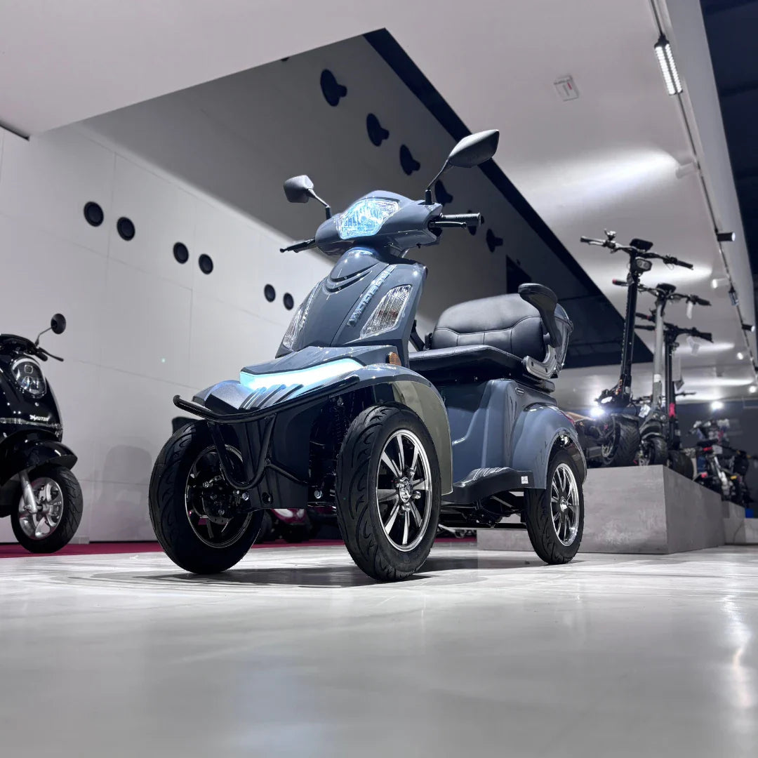 Izdelek na sliki je električni mobilni skuter, model Vigorous Vista Q1. Skuter ima štiri kolesa, kar omogoča stabilnejšo vožnjo. Na sliki je vidna moderna zasnova z udobnim sedežem in svetlobnimi elementi na sprednjem delu. V ozadju so prikazani tudi drugi električni skuterji, kar nakazuje na razstavo ali trgovino z električnimi vozili.