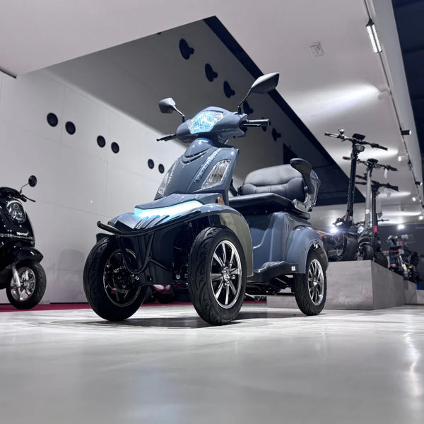 Izdelek na sliki je električni mobilni skuter, model Vigorous Vista Q1. Skuter ima štiri kolesa, kar omogoča stabilnejšo vožnjo. Na sliki je vidna moderna zasnova z udobnim sedežem in svetlobnimi elementi na sprednjem delu. V ozadju so prikazani tudi drugi električni skuterji, kar nakazuje na razstavo ali trgovino z električnimi vozili.