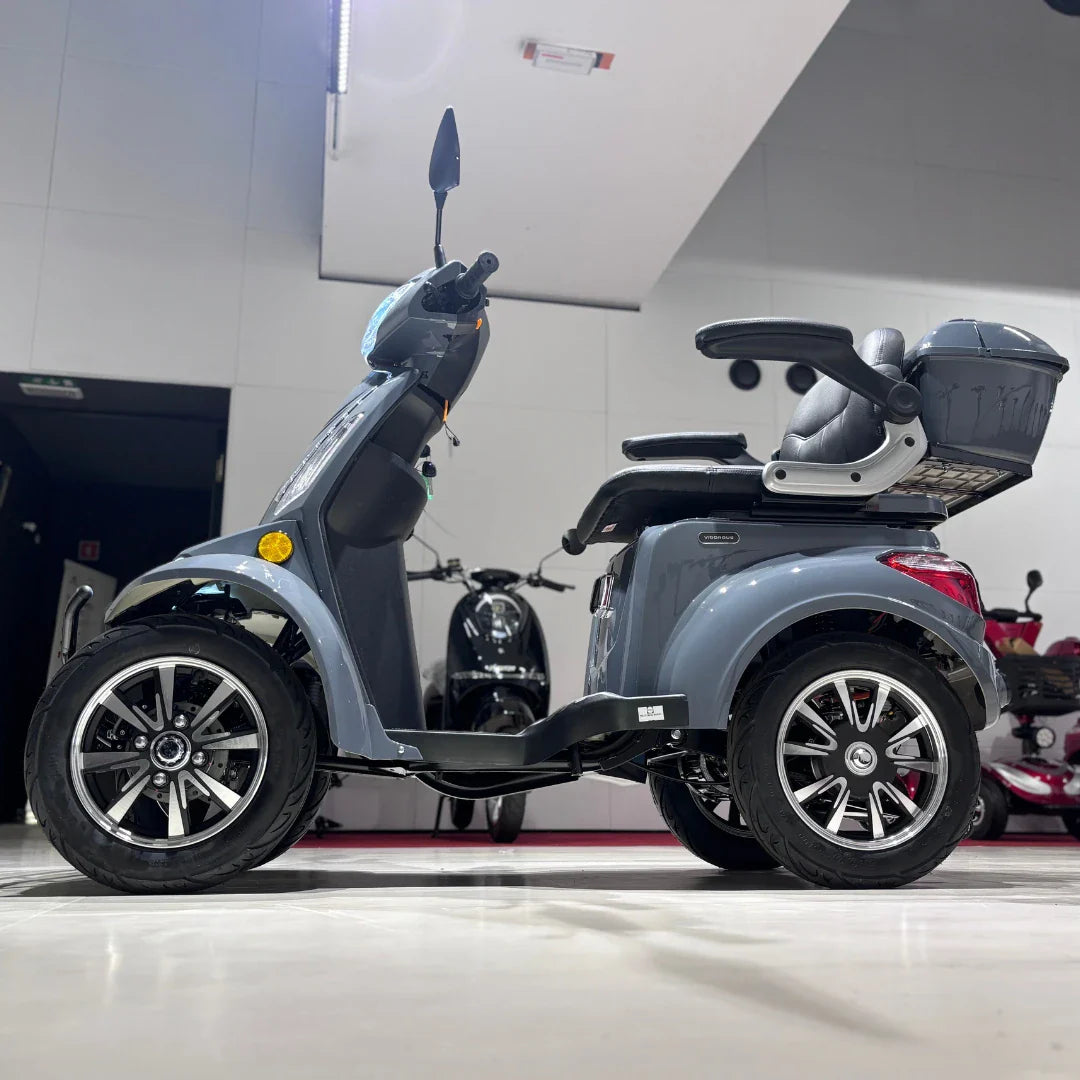 Izdelek je električni mobilni skuter, model Vigorous Vista Q1. Na sliki je prikazan skuter s štirimi kolesi, ki ima udobno sedežno mesto in prostoren prtljažnik. Barva skuterja je siva, opremljen je z modernimi kolesi in ima vgrajene svetilke ter ogledala. V ozadju so vidni tudi drugi podobni skuterji.