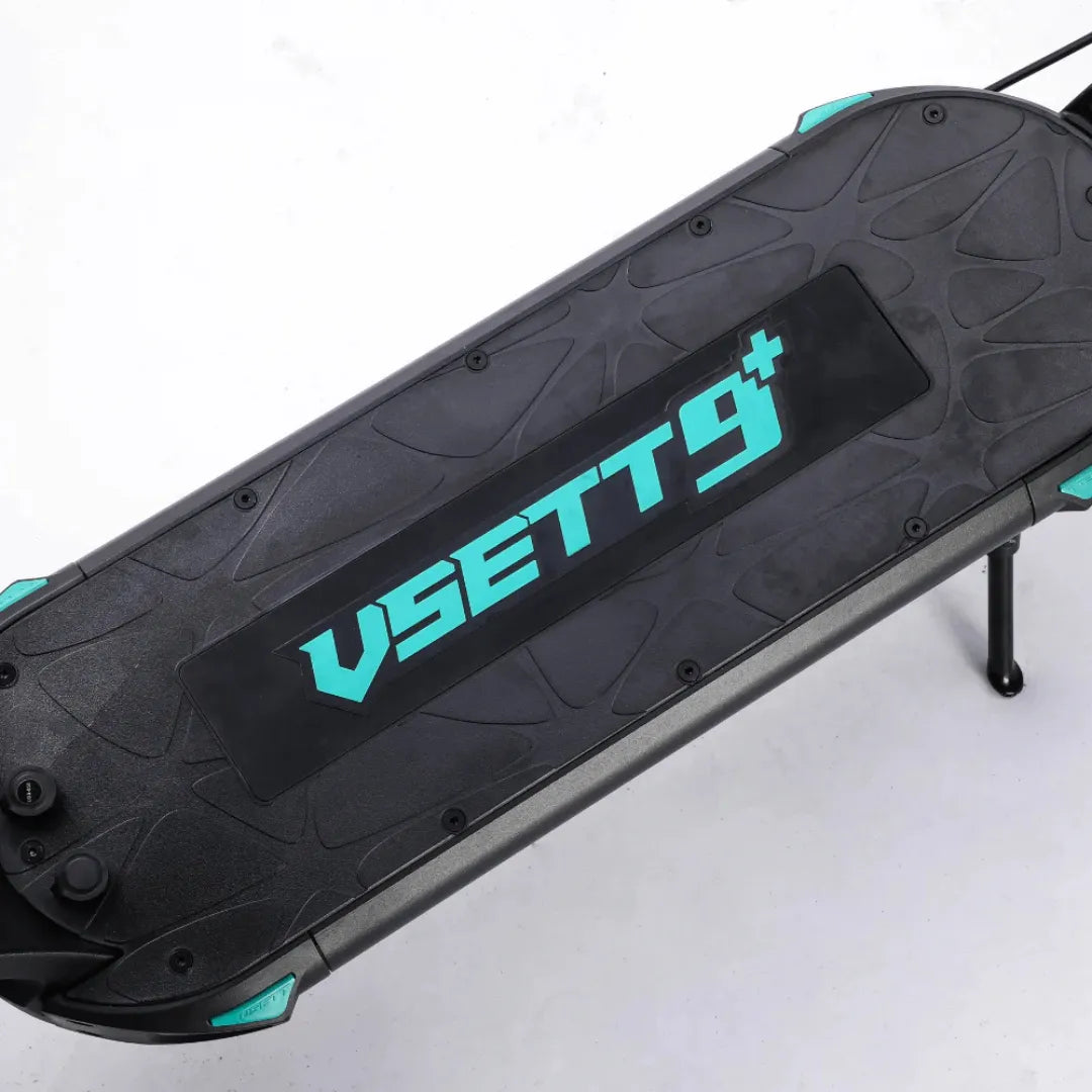 Izdelek na sliki je električni skiro, znamke Vsett, model Vsett 9. Na sliki je vidna platforma skiroja, ki ima črno površino z modrimi detajli in logotipom Vsett 9. Vidni so tudi elementi za pritrditev in podrobnosti na robovih, kar nakazuje na robustno zasnovo skiroja.