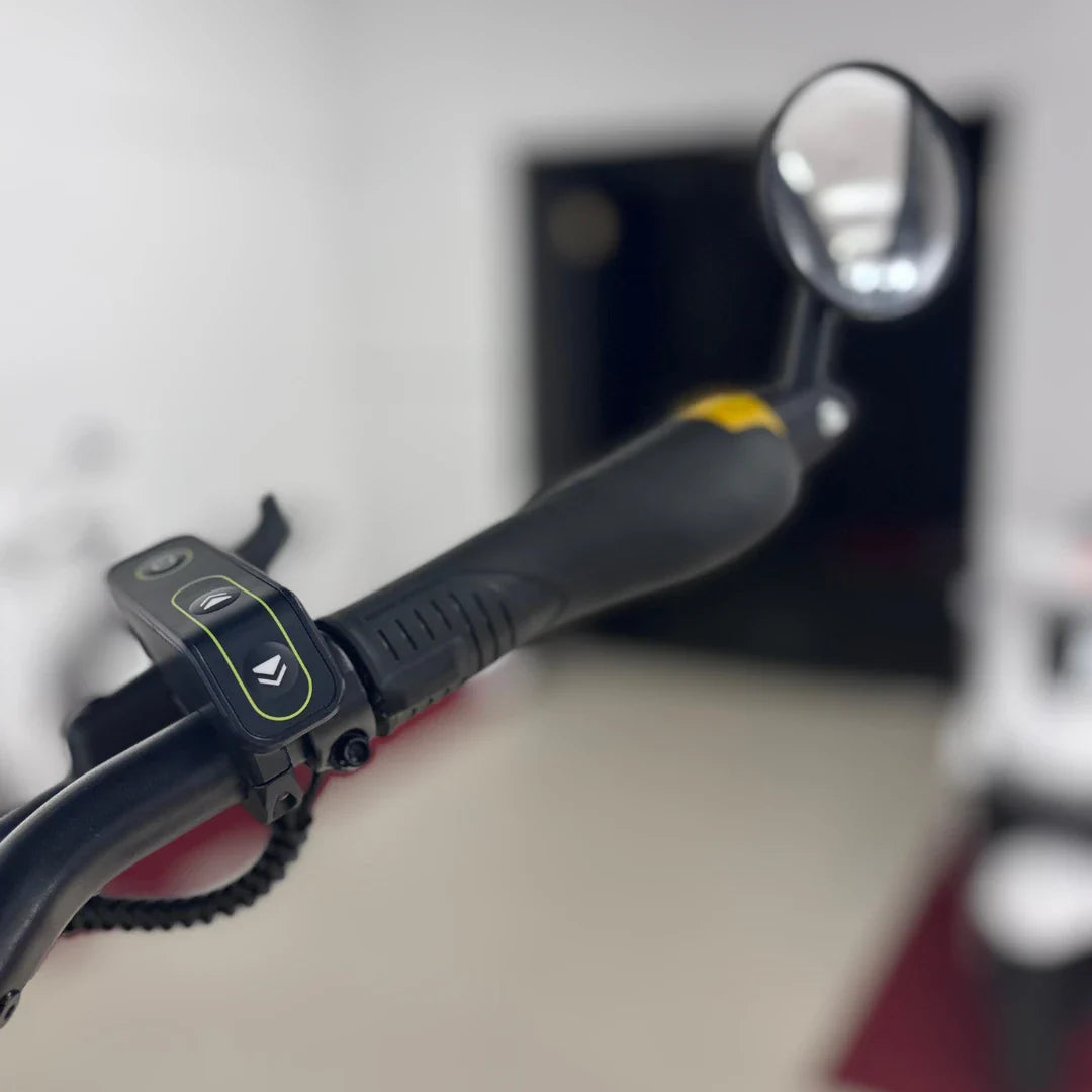 Na sliki je električni skiro znamke Vsett, model 11 Super. Vidna je ročica z gumbi za upravljanje, ki omogočajo prilagoditev hitrosti in delovanje luči. Ročaj je zasnovan za udobno držanje, kar je pomembno za varno vožnjo. V ozadju je nejasen prostor, kar daje občutek, da je skiro pripravljen za uporabo.