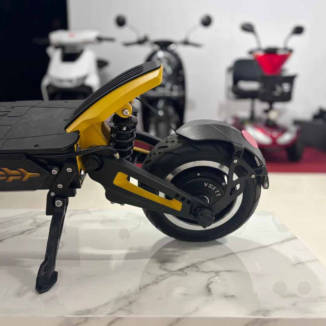 Na sliki je prikazan električni skiro, model Vsett 11 Super. Vidna je zadnja stran skiroja z močnim kolesom in vzmetenjem, kar nakazuje na udobno vožnjo. Kolo ima značilen dizajn z oznako "VSETT", kar potrjuje znamko izdelka. V ozadju so vidni tudi drugi električni prevozi, kar daje občutek o sodobni mobilnosti.