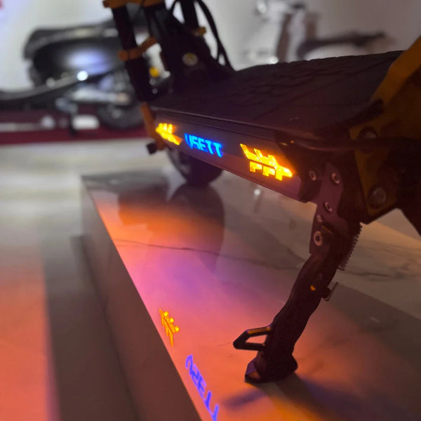 Na sliki je električni skiro znamke VSETT, model 11 Super. Vidna je podrobnost skiroja, vključno z osvetlitvijo, ki je nameščena na stranski strani. Osvetlitev je v različnih barvah, kar daje skiroju sodoben in privlačen videz. Na sliki je tudi del podnožja skiroja ter stojalo, ki omogoča stabilno parkiranje. Ozadje je nejasno, kar poudari glavni izdelek.