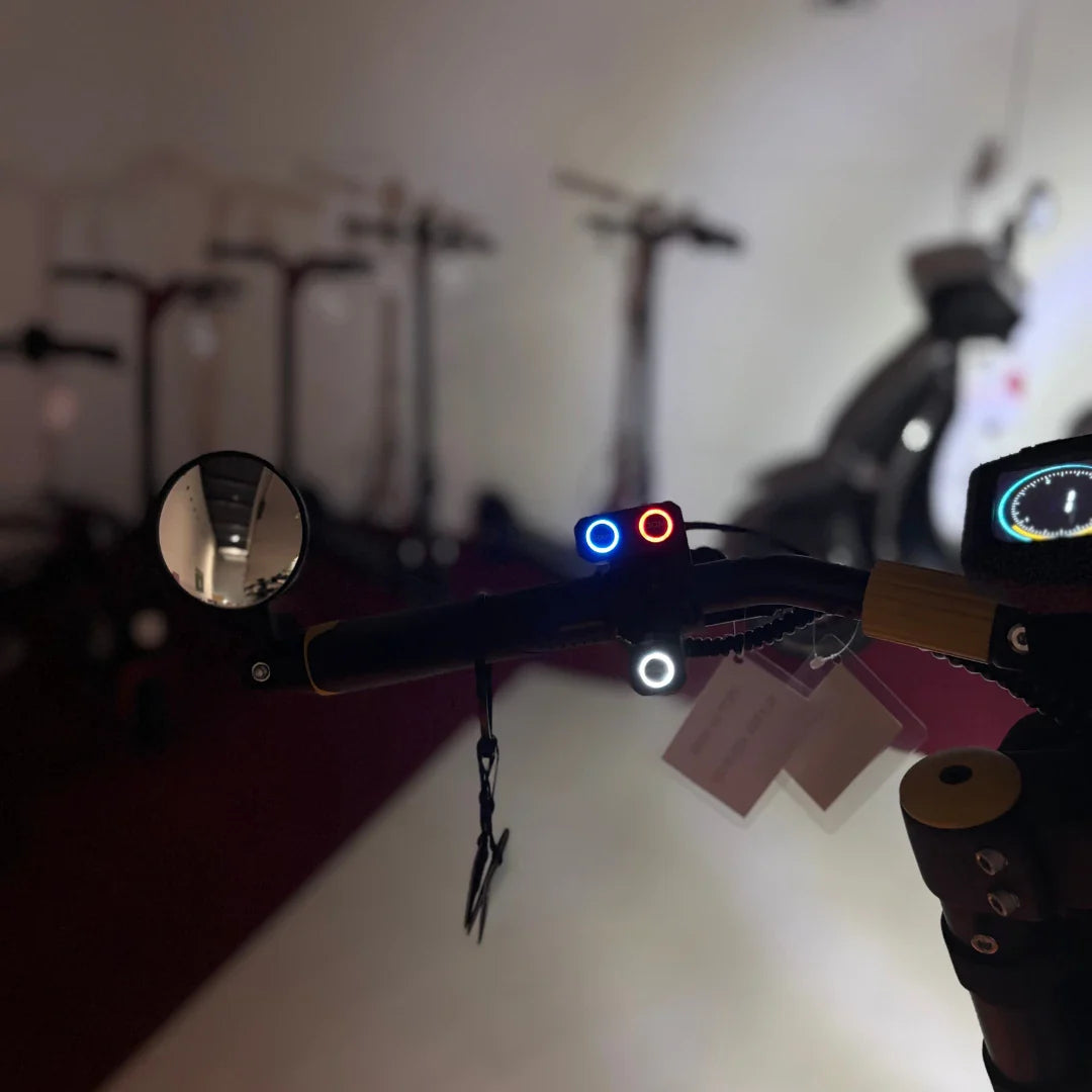 Na sliki je električni skiro znamke Vsett, model 11 Super. Vidna je sprednja stran krmila, kjer so nameščeni luči v rdeči in modri barvi ter ogledalo za boljšo vidljivost. V ozadju so drugi skiroji, kar nakazuje, da gre za prodajni prostor.