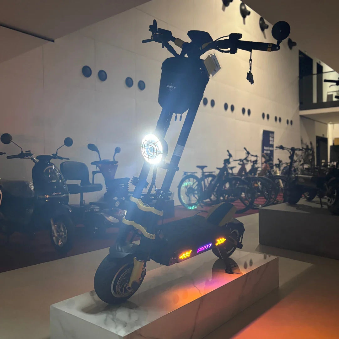 Na sliki je električni skiro znamke Vsett, model 11 Super. Izdelek je postavljen na podstavek in je osvetljen, kar poudarja njegove svetlobne elemente. Na skiroju so vidni široki pnevmatiki, robusten okvir ter LED luči, ki dodajajo sodoben videz. V ozadju so vidni tudi drugi električni skiroji in kolesa, kar nakazuje na razstavo električnih prevoznih sredstev.