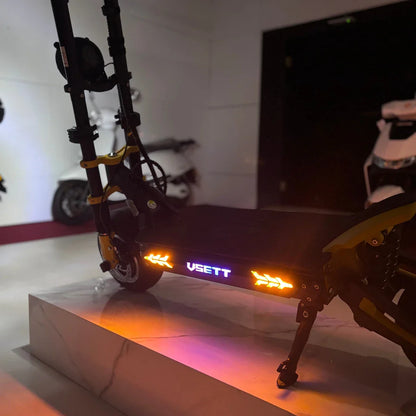 Na sliki je električni skiro znamke VSETT, model 11 Super. Vidna je njegova robustna konstrukcija, z osvetlitvijo na platformi, ki prikazuje ime znamke. Skuter ima tudi vzmetenje in je postavljen na svetli površini, kar poudarja njegov sodoben dizajn. V ozadju so vidne druge prevozne naprave.