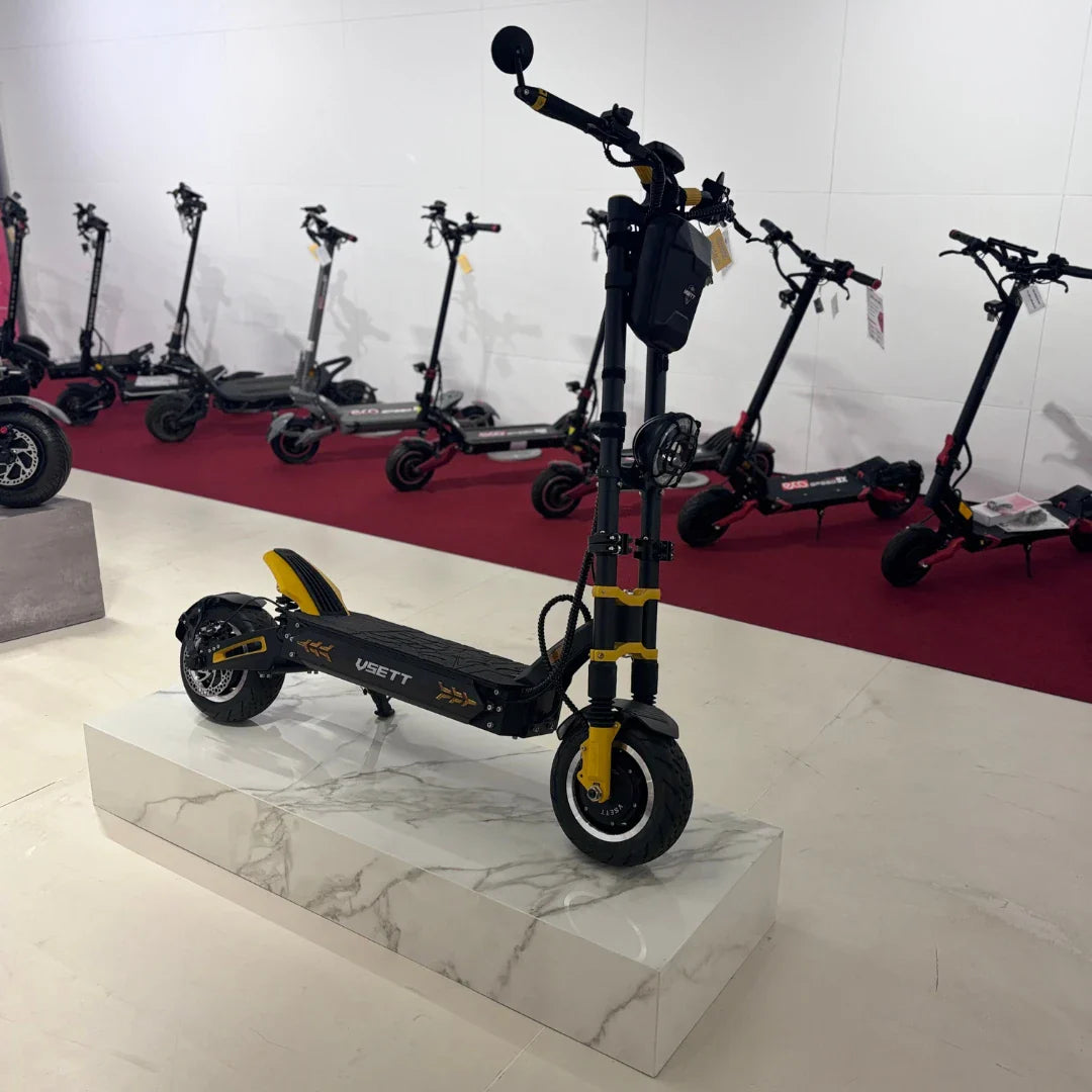 Na sliki je električni skiro znamke Vsett, model 11 Super. Izdelek je postavljen na svetel podstavek, kar izpostavlja njegov športni dizajn. Na skiroju so vidne široke pnevmatike in robustna konstrukcija, kar nakazuje na dobro stabilnost in zmogljivost. V ozadju so vidni tudi drugi električni skiroji, kar nakazuje, da gre za razstavo ali prodajno mesto.