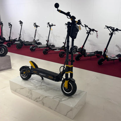 Na sliki je električni skiro znamke Vsett, model 11 Super. Izdelek je postavljen na svetel podstavek, kar izpostavlja njegov športni dizajn. Na skiroju so vidne široke pnevmatike in robustna konstrukcija, kar nakazuje na dobro stabilnost in zmogljivost. V ozadju so vidni tudi drugi električni skiroji, kar nakazuje, da gre za razstavo ali prodajno mesto.