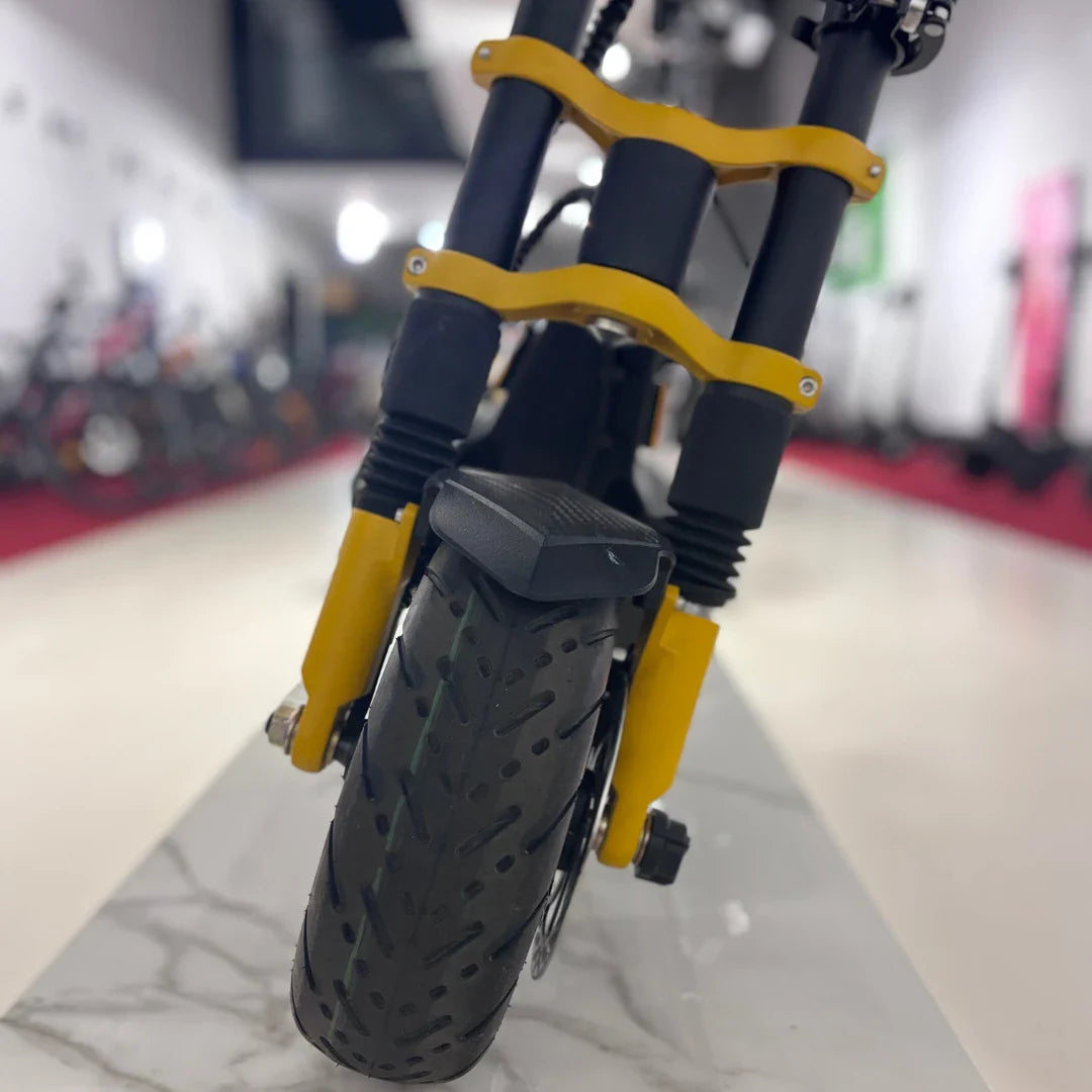 Na sliki je električni skiro, model Vsett 11 Super. Vidna je sprednja stran skiroja, ki vključuje robustne vzmeti in široko pnevmatiko z vzorcem, kar nakazuje na dobro oprijemljivost. Zasnova je moderna, z rumenimi in črnimi elementi, kar daje skiroju dinamičen videz. V ozadju so vidni tudi drugi skiroji, kar nakazuje, da gre za prodajno mesto.