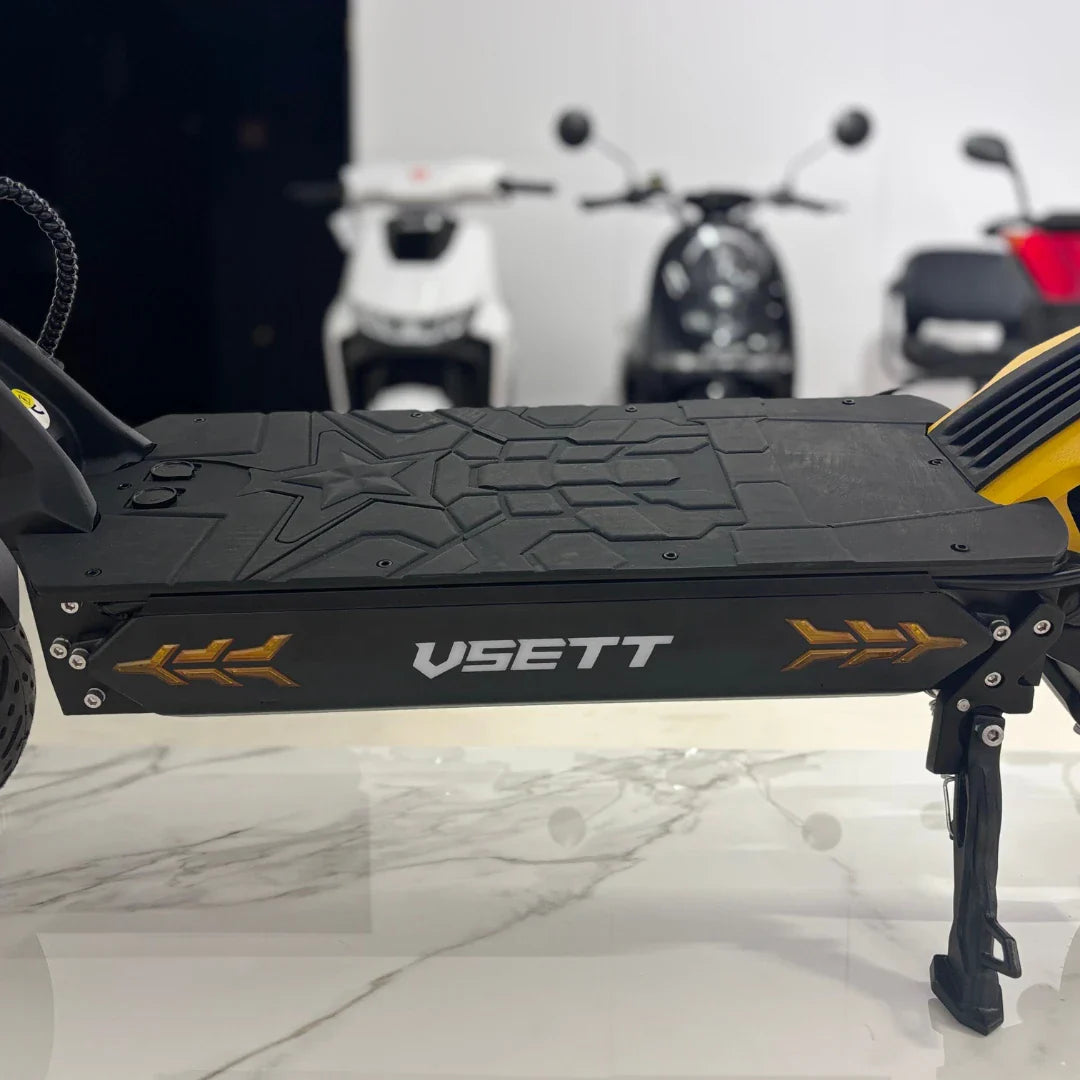 Na sliki je električni skiro znamke VSETT, model 11 Super. Vidna je platforma skiroja z značilnim vzorcem na površini za stopala ter logotipom VSETT na strani. V ozadju so delno vidni drugi skiroji, kar nakazuje, da je slika posneta v trgovini ali razstavnem prostoru.