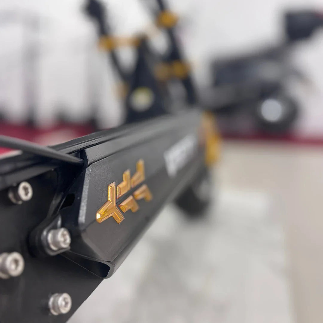 Na sliki je električni skiro, model Vsett 11 Super. Vidna je črna konstrukcija skiroja z zlatimi detajli, ki poudarjajo njegov športni videz. Na sliki so tudi vidni nekateri vijaki in del okvirja, kar nakazuje na robustno zasnovo izdelka. Ozadje je zamegljeno, kar omogoča osredotočanje na detajle skiroja.