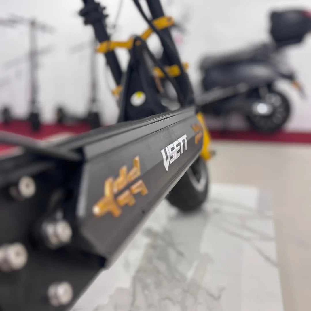 Na sliki je električni skiro znamke Vsett, model 11 Super. Vidna je platforma skiroja s poudarjenim logotipom Vsett, ki je v zlati barvi. V ozadju so vidni drugi električni skiroji, kar nakazuje, da gre za razstavo ali trgovino z električnimi vozili.