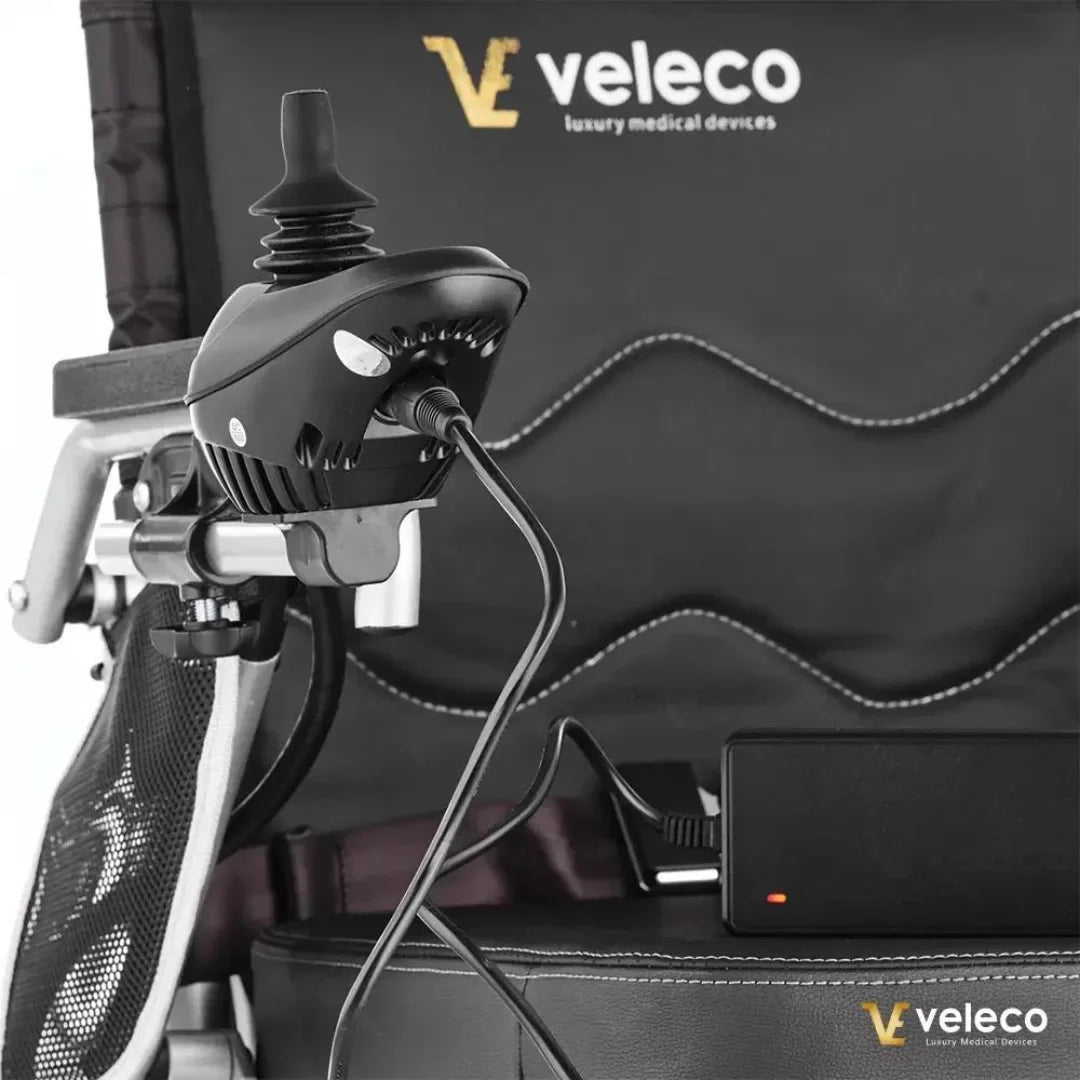 Izdelek na sliki je električni invalidski voziček znamke Veleco, model Cosmo. Na sliki je prikazan del upravljalnika vozička, ki ima ergonomsko obliko in je povezan z napajalnikom. V ozadju je vidna tudi oblazinjena površina vozička, kar nakazuje na udobje pri uporabi.