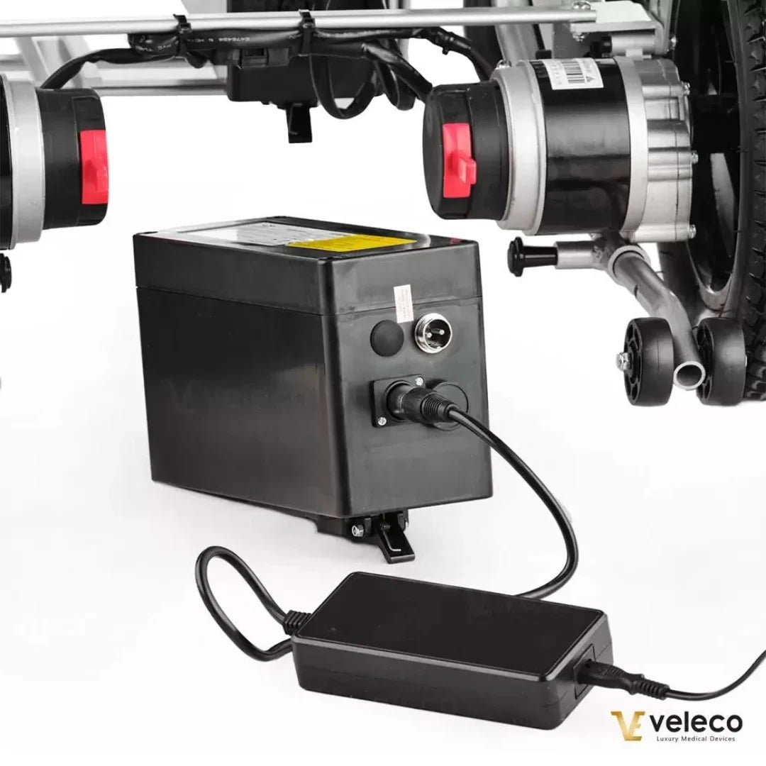 Izdelek je električni invalidski voziček, znamke Veleco, model Cosmo. Na sliki je prikazana baterija vozička, ki je povezana z električnim motorjem, ter polnilnik. Baterija je črne barve in ima vtičnico za povezavo, kar omogoča enostavno polnjenje. Vidna sta tudi dva motorja, ki sta nameščena ob kolesih.