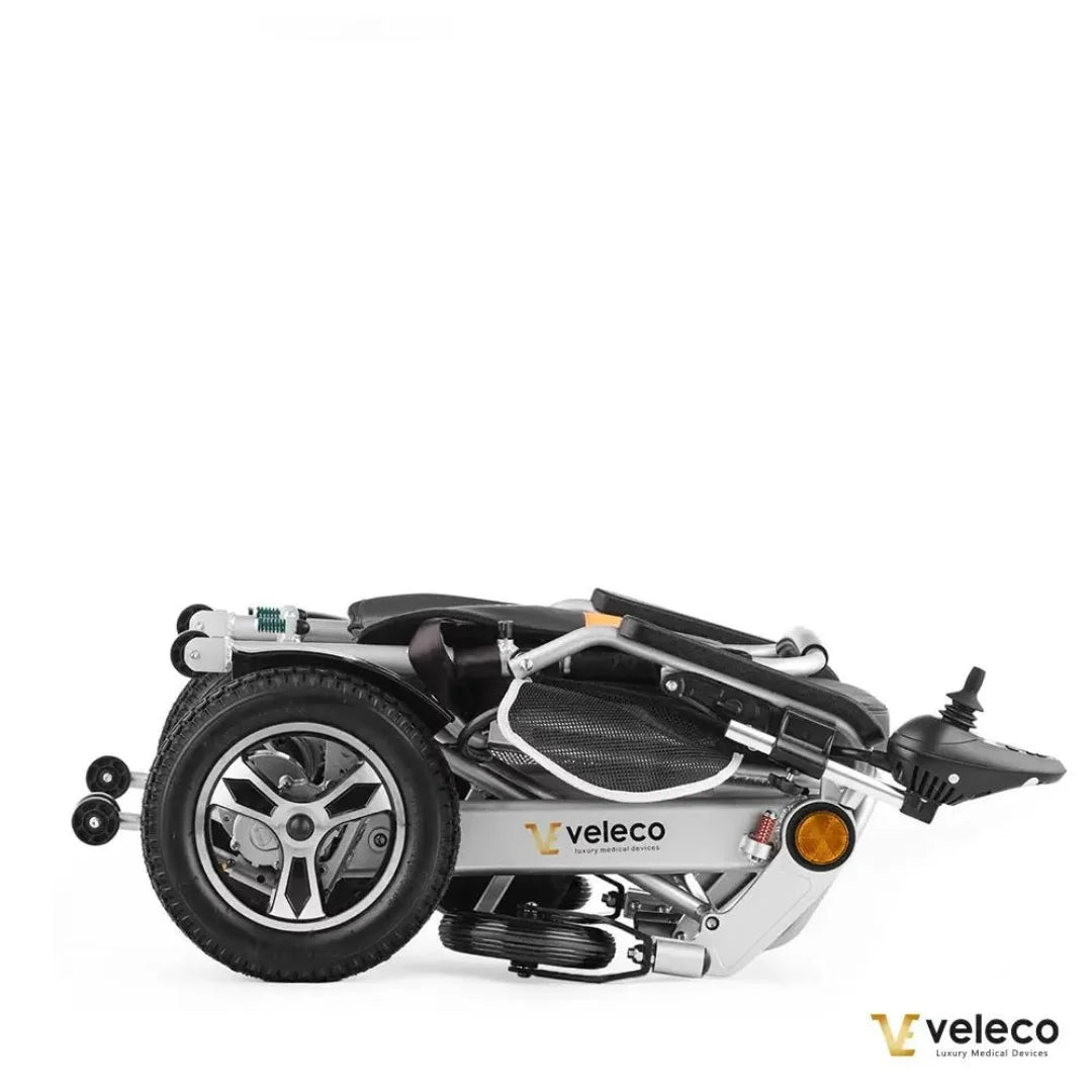 Izdelek je električni invalidski voziček znamke Veleco, model Cosmo. Na sliki je prikazan zložen voziček, ki ima kompaktno obliko in je opremljen z manjšimi kolesi ter udobnim sedežem. Vidna je tudi kontrolna enota, ki omogoča enostavno upravljanje vozička.
