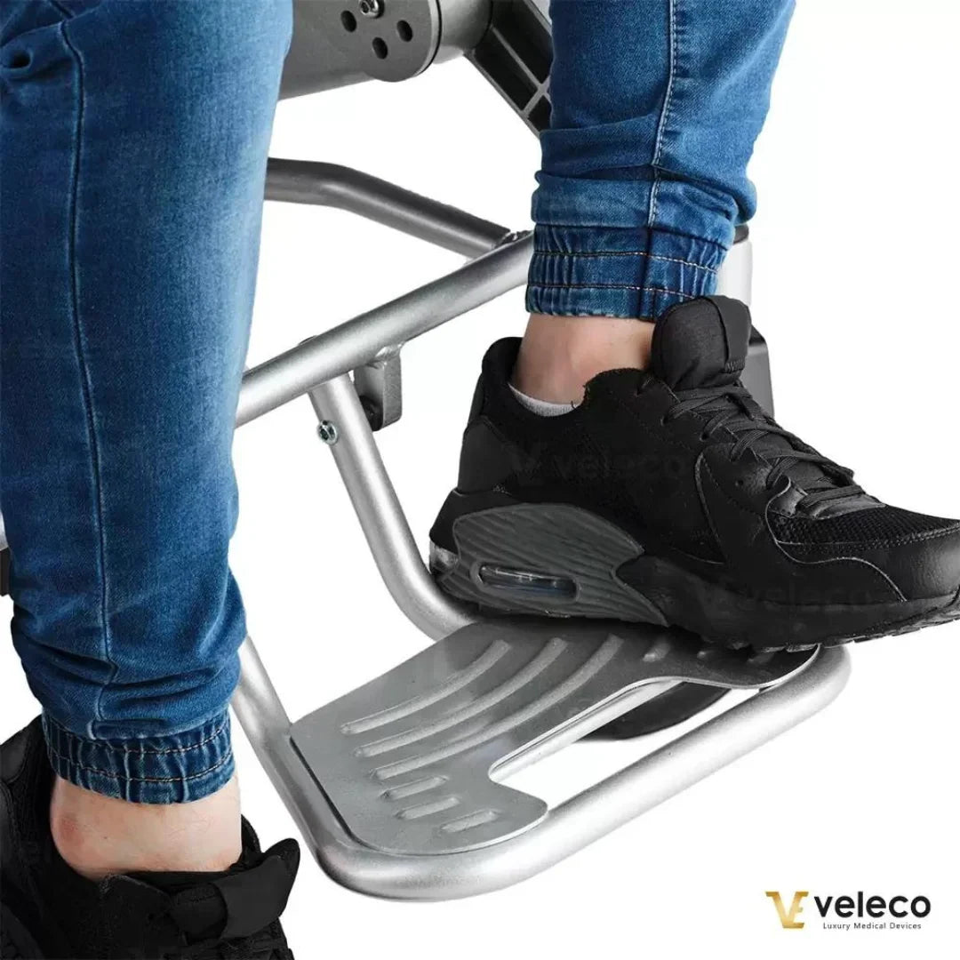 Izdelek na sliki je električni invalidski voziček znamke Veleco, model Cosmo. Na sliki so vidni nogi uporabnika, ki stojita na aluminijastem podnožju vozička, oblečeni v modre jeans hlače in črne športne copate. Voziček je zasnovan za udobno in varno premikanje uporabnika.