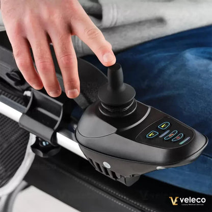 Izdelek je električni invalidski voziček znamke Veleco, model Cosmo. Na sliki je prikazan del upravljalne palice, ki jo uporablja uporabnik, ter gumbi za upravljanje vozička. Vidna je roka, ki se dotika palice, kar nakazuje na aktivno uporabo vozička. Ozadje prikazuje del oblačil in sedeža vozička.