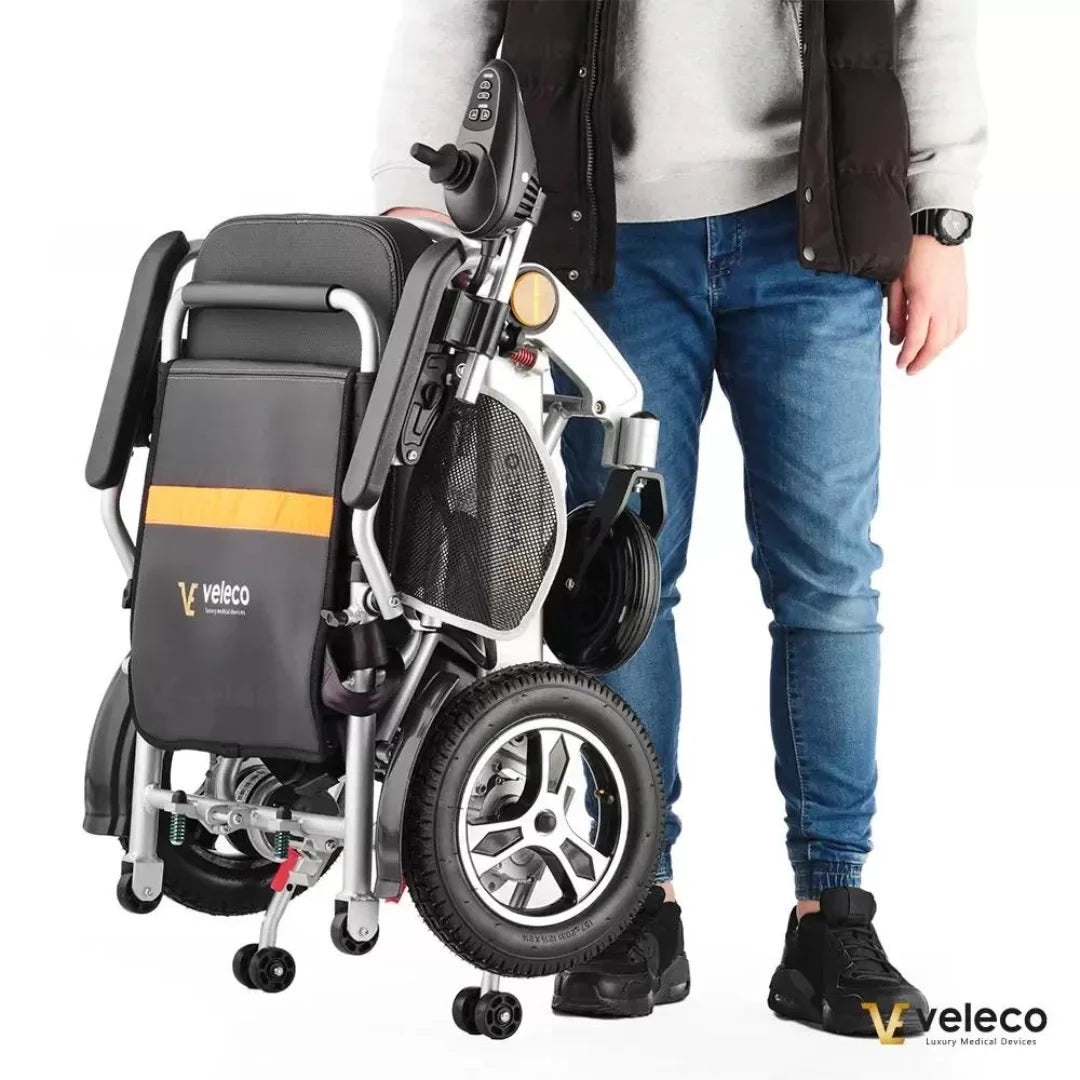 Izdelek na sliki je električni invalidski voziček znamke Veleco, model Cosmo. Na sliki je videti voziček v zloženem stanju, kar omogoča enostavno prenašanje. Ob njem stoji oseba, ki ga drži za ročaj, kar nakazuje na njegovo prenosljivost in praktičnost. Voziček ima sodoben dizajn in je opremljen z manjšimi kolesi, kar olajša premikanje.