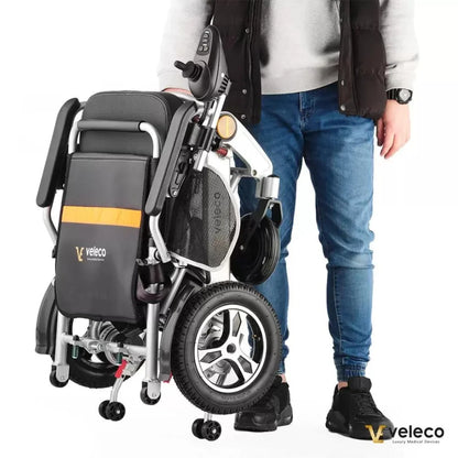 Izdelek na sliki je električni invalidski voziček znamke Veleco, model Cosmo. Na sliki je videti voziček v zloženem stanju, kar omogoča enostavno prenašanje. Ob njem stoji oseba, ki ga drži za ročaj, kar nakazuje na njegovo prenosljivost in praktičnost. Voziček ima sodoben dizajn in je opremljen z manjšimi kolesi, kar olajša premikanje.