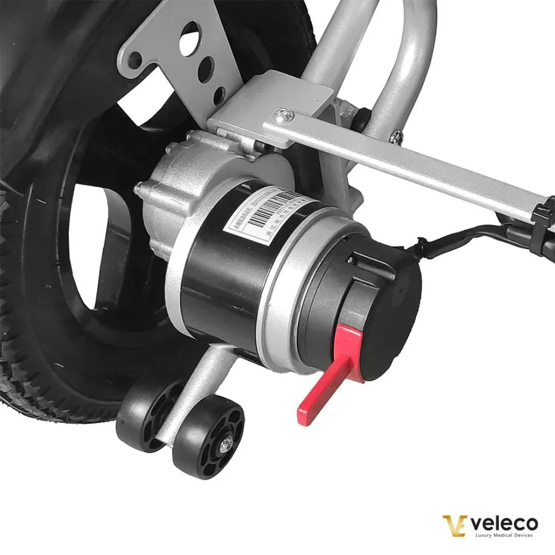 Izdelek na sliki je električni invalidski voziček znamke Veleco, model Cosmo. Na sliki je prikazan del motorja, ki je nameščen na zadnjem kolesu vozička. Motor je v kovinskem ohišju, obdan z mehanizmom za pritrditev, ter ima rdečo ročico, ki verjetno služi za upravljanje ali nastavitev. Kolo je opremljeno z gumijastim profilom, kar omogoča dobro oprijemljivost na različnih površinah.