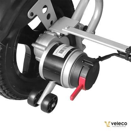 Izdelek na sliki je električni invalidski voziček znamke Veleco, model Cosmo. Na sliki je prikazan del motorja, ki je nameščen na zadnjem kolesu vozička. Motor je v kovinskem ohišju, obdan z mehanizmom za pritrditev, ter ima rdečo ročico, ki verjetno služi za upravljanje ali nastavitev. Kolo je opremljeno z gumijastim profilom, kar omogoča dobro oprijemljivost na različnih površinah.