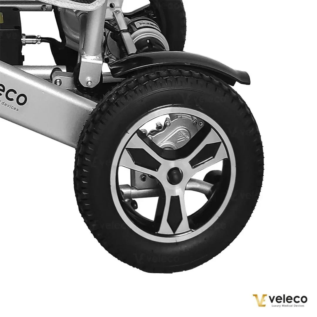 Izdelek na sliki je električni invalidski voziček, model Veleco Cosmo. Na sliki je prikazan del vozička, osredotočen na kolo, ki ima sodoben dizajn z aluminijastimi platišči in širokimi pnevmatikami, kar omogoča dobro stabilnost in udobje pri vožnji. Vidna je tudi struktura okvirja, ki je izdelana iz trpežnih materialov.